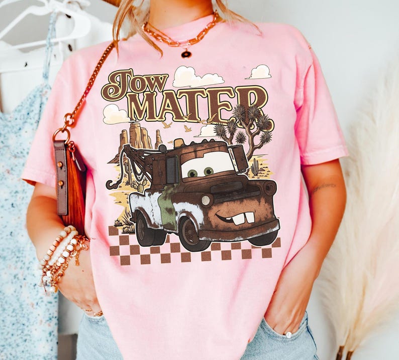 May include: A light pink t-shirt featuring a graphic of the character Tow Mater from the movie Cars. The design includes the text Tow Mater, a desert landscape with cacti, and a brown and white checkered pattern along the bottom edge.