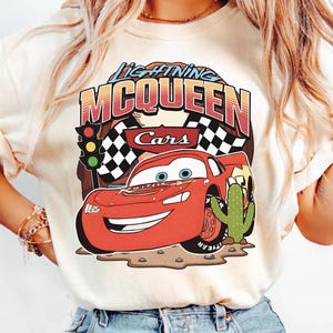 May include: A cream-coloured t-shirt featuring a graphic of the red race car Lightning McQueen from the movie Cars. The design includes the text Lightning McQueen, a traffic light, checkered flags, and a green cactus on a brown desert ground.