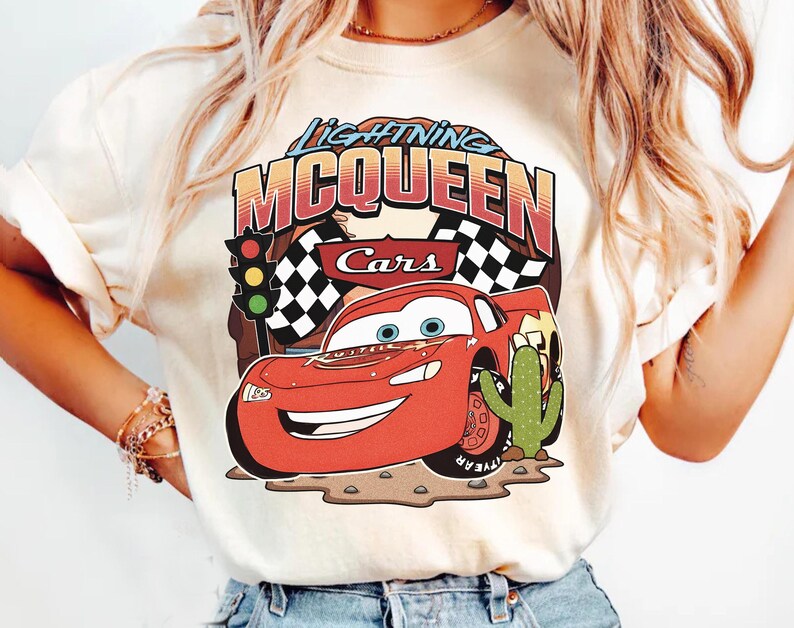 May include: A cream-coloured t-shirt featuring a graphic of the red race car Lightning McQueen from the movie Cars. The design includes the text Lightning McQueen, a traffic light, checkered flags, and a green cactus on a brown desert ground.
