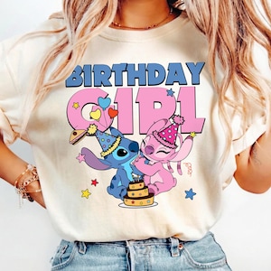 May include: Cream-colored t-shirt featuring the text "BIRTHDAY GIRL" in blue and pink. The design includes cartoon characters Stitch and Angel, wearing party hats, with a cake and stars.
