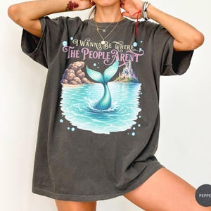 K&ouml;nnte beinhalten: Ein anthrazitfarbenes T-Shirt mit einem Grafikdruck einer Meerjungfrauenflosse, die in blauem Wasser spritzt, einem felsigen Ufer und einem Schloss. Der Text lautet, I WANNA BE WHERE THE PEOPLE AREN'T. Das T-Shirt hat kurze &Auml;rmel und einen Rundhalsausschnitt.
