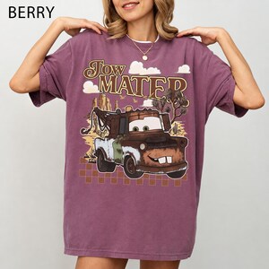 May include: A berry-colored short-sleeve t-shirt featuring a graphic print of the character Tow Mater. The design includes the text "Tow Mater" above the truck, set against a desert landscape with clouds, cacti, and a checkered pattern base.