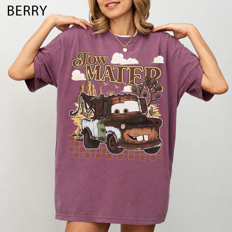 May include: A berry-colored short-sleeve t-shirt featuring a graphic print of the character Tow Mater. The design includes the text "Tow Mater" above the truck, set against a desert landscape with clouds, cacti, and a checkered pattern base.