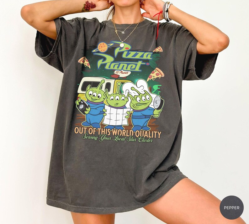 May include: A charcoal gray short-sleeve t-shirt featuring a Pizza Planet graphic with three green aliens, a yellow delivery truck, and pizza slices. The text reads Pizza Planet, Out of This World Quality, and Serving Your Local Star Cluster.