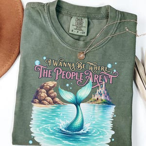 K&ouml;nnte beinhalten: Ein olivgr&uuml;nes T-Shirt mit einer Grafik einer Meerjungfrauenflosse, die in blauem Wasser spritzt, einer Felsformation und einem Schloss. Der Text lautet "I WANNA BE WHERE THE PEOPLE AREN'T" in rosa und goldener Schrift. Rundhalsausschnitt und Etikett.