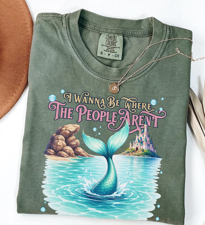 K&ouml;nnte beinhalten: Ein olivgr&uuml;nes T-Shirt mit einer Grafik einer Meerjungfrauenflosse, die in blauem Wasser spritzt, einer Felsformation und einem Schloss. Der Text lautet "I WANNA BE WHERE THE PEOPLE AREN'T" in rosa und goldener Schrift. Rundhalsausschnitt und Etikett.