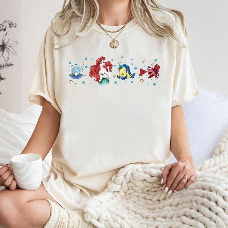 Może przedstawiać: Kremowy t-shirt z kr&oacute;tkim rękawem z nadrukiem graficznym na piersi przedstawiającym niebieską muszlę z perłą, rudowłosą syrenę, ż&oacute;łto-niebieską rybę oraz czerwonego kraba, otoczonych małymi niebieskimi kropkami i ż&oacute;łtymi gwiazdkami.
