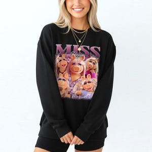 May include: Black sweatshirt featuring a graphic of Miss Piggy and the word "MISS" in a purple, stylized font. The graphic includes multiple images of Miss Piggy. The sweatshirt is worn by a person.
