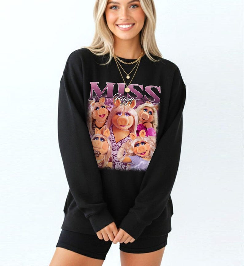 May include: Black sweatshirt featuring a graphic of Miss Piggy and the word "MISS" in a purple, stylized font. The graphic includes multiple images of Miss Piggy. The sweatshirt is worn by a person.