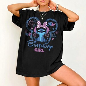 May include: Black t-shirt featuring a cartoon character with a purple bow and the text "Birthday Girl" in blue and pink. The design includes a Mickey Mouse ear shape with fireworks and floral accents.