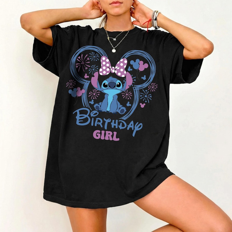 May include: Black t-shirt featuring a cartoon character with a purple bow and the text "Birthday Girl" in blue and pink. The design includes a Mickey Mouse ear shape with fireworks and floral accents.