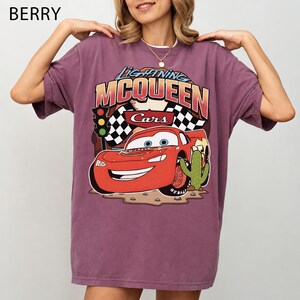 May include: A berry-coloured short-sleeve t-shirt featuring a graphic print of Lightning McQueen from the movie Cars. The design includes the text Lightning McQueen, a chequered flag, a traffic light, and a cactus on a desert-like ground.