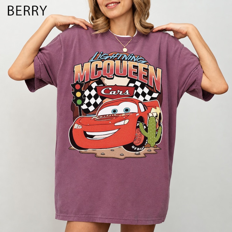 May include: A berry-coloured short-sleeve t-shirt featuring a graphic print of Lightning McQueen from the movie Cars. The design includes the text Lightning McQueen, a chequered flag, a traffic light, and a cactus on a desert-like ground.