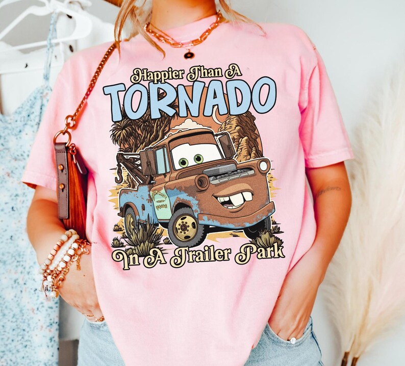 Op de afbeelding: Een lichtroze t-shirt met een afbeelding van een roestige bruine en blauwe takelwagen met grote ogen en een brede glimlach. De tekst erboven luidt Happier Than A TORNADO en de tekst eronder In A Trailer Park in een gestileerd lettertype.