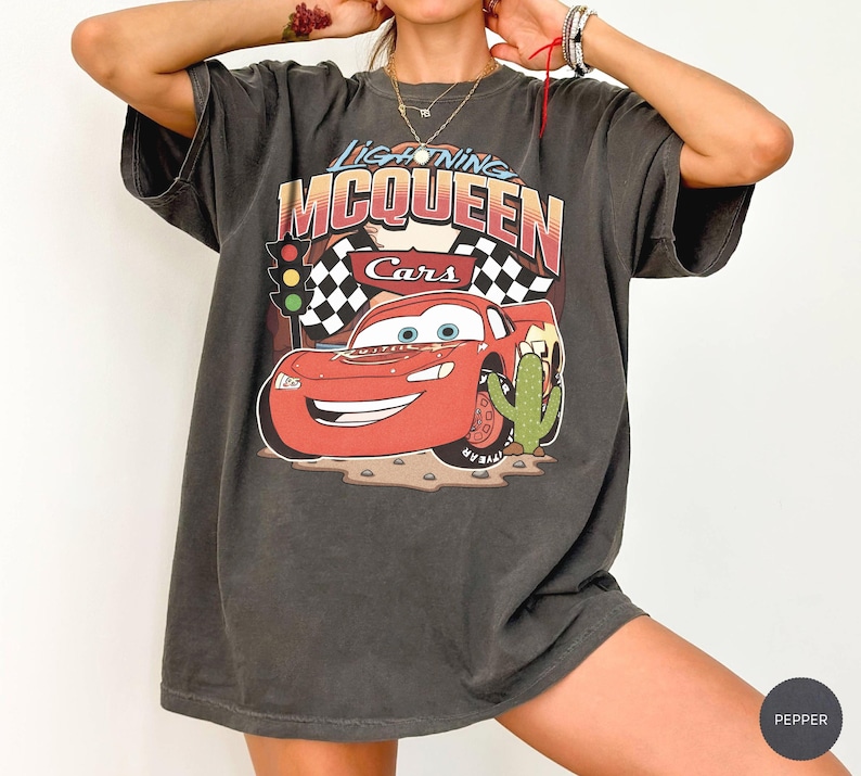 May include: A charcoal grey short-sleeved t-shirt featuring a graphic print of the character Lightning McQueen from the film Cars. The design includes the text Lightning McQueen, a traffic light, checkered flags, and a cactus illustration.