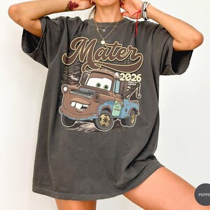 May include: A charcoal gray short-sleeve t-shirt featuring a graphic print of the character Mater from the movie Cars. The design includes the text Mater and 2026 in a retro font, with a brown and blue tow truck illustration set against a forest.