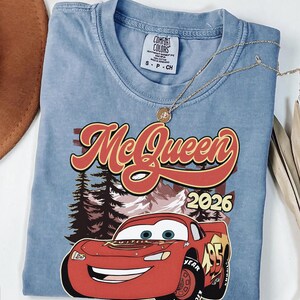 Puede incluir: Una camiseta azul de manga corta con un gr&aacute;fico de un coche de carreras rojo con el n&uacute;mero 95, el texto McQueen y 2026, y un fondo de bosque y monta&ntilde;as. La camiseta tiene una etiqueta de la marca Comfort Colors y un collar dorado.