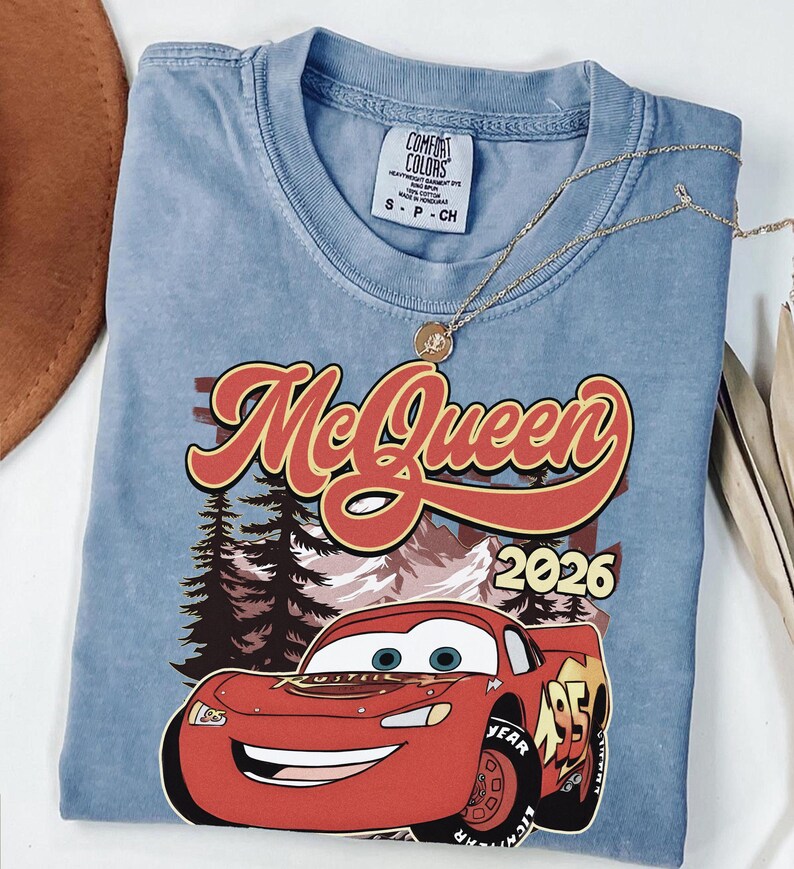 Puede incluir: Una camiseta azul de manga corta con un gr&aacute;fico de un coche de carreras rojo con el n&uacute;mero 95, el texto McQueen y 2026, y un fondo de bosque y monta&ntilde;as. La camiseta tiene una etiqueta de la marca Comfort Colors y un collar dorado.