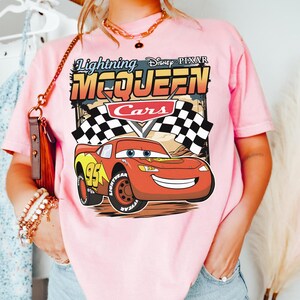 May include: A light pink t-shirt featuring a graphic print of the character Lightning McQueen from the Disney Pixar movie Cars. The design includes the text Lightning McQueen, the Cars logo, and checkered flags against a desert background.