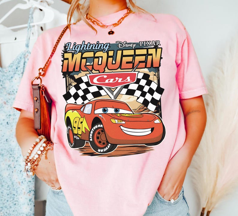 May include: A light pink t-shirt featuring a graphic print of the character Lightning McQueen from the Disney Pixar movie Cars. The design includes the text Lightning McQueen, the Cars logo, and checkered flags against a desert background.