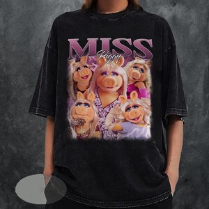 May include: Black t-shirt featuring a graphic of Miss Piggy and the text "MISS Piggy" in a pink font. The image shows Miss Piggy in various poses. The shirt has a vintage, washed-out look.