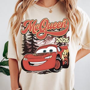Puede incluir: Una camiseta de manga corta de color crema con un estampado gr&aacute;fico de un coche de carreras rojo con el n&uacute;mero 95. Sobre el coche, el texto McQueen est&aacute; escrito en una fuente roja estilizada, con el a&ntilde;o 2026 a la derecha del paisaje forestal.