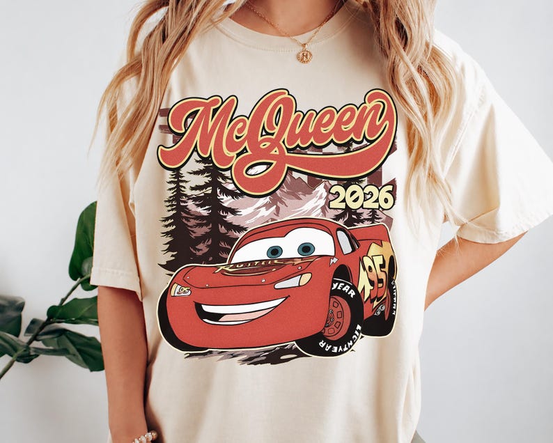 Puede incluir: Una camiseta de manga corta de color crema con un estampado gr&aacute;fico de un coche de carreras rojo con el n&uacute;mero 95. Sobre el coche, el texto McQueen est&aacute; escrito en una fuente roja estilizada, con el a&ntilde;o 2026 a la derecha del paisaje forestal.