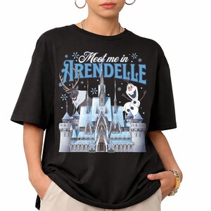May include: A black short-sleeved t-shirt featuring a graphic print of an ice castle, a reindeer, and a snowman. The text "Meet me in ARENDELLE" is printed in blue and white above the castle, surrounded by small white snowflake illustrations.