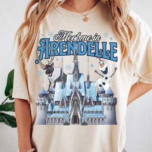 May include: A cream-coloured t-shirt featuring a graphic print of an ice castle, a reindeer, and a snowman. The text "Meet me in ARENDELLE" is printed in blue above the castle. The design includes light blue snowflake accents throughout the graphic.