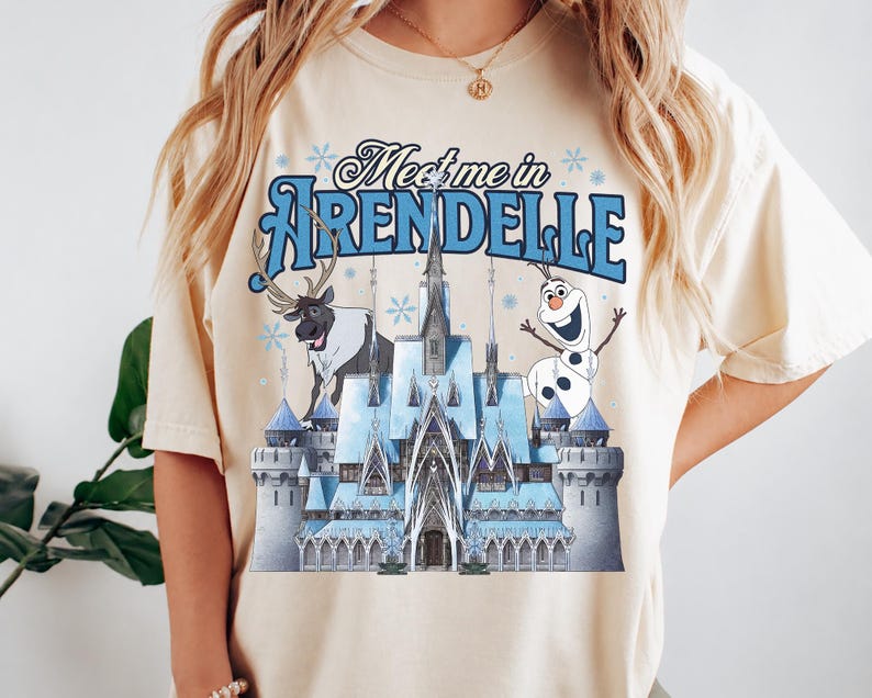 May include: A cream-coloured t-shirt featuring a graphic print of an ice castle, a reindeer, and a snowman. The text "Meet me in ARENDELLE" is printed in blue above the castle. The design includes light blue snowflake accents throughout the graphic.