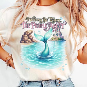 K&ouml;nnte beinhalten: Ein cremefarbenes T-Shirt mit einem Grafikdruck einer t&uuml;rkisfarbenen Meerjungfrauenflosse, die aus blauem Wasser ragt, sowie einer felsigen K&uuml;ste, einem Schloss auf einer Insel und Blasen. Der Text lautet I WANNA BE WHERE THE PEOPLE ARENT.