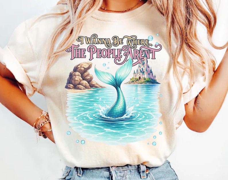 K&ouml;nnte beinhalten: Ein cremefarbenes T-Shirt mit einem Grafikdruck einer t&uuml;rkisfarbenen Meerjungfrauenflosse, die aus blauem Wasser ragt, sowie einer felsigen K&uuml;ste, einem Schloss auf einer Insel und Blasen. Der Text lautet I WANNA BE WHERE THE PEOPLE ARENT.