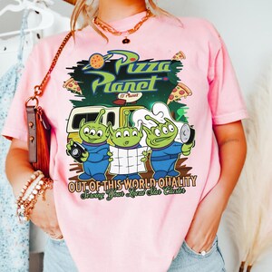 May include: A light pink t-shirt featuring a graphic of three green three-eyed aliens in blue uniforms. The text reads Pizza Planet, Out of This World Quality, and Serving Your Local Star Cluster. The design includes a yellow delivery truck and pizza slices.