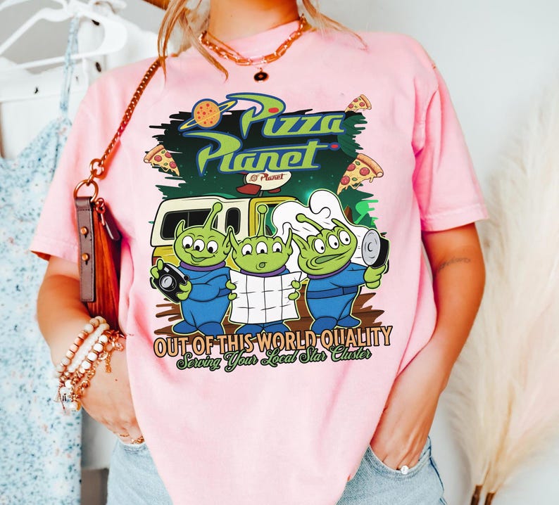 May include: A light pink t-shirt featuring a graphic of three green three-eyed aliens in blue uniforms. The text reads Pizza Planet, Out of This World Quality, and Serving Your Local Star Cluster. The design includes a yellow delivery truck and pizza slices.