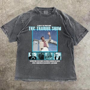 Pode incluir: T-shirt cinzento escuro com um design gráfico do filme "The Truman Show". O design inclui o título do filme, uma cena do filme e uma citação de Joan Didion. A t-shirt tem gola redonda e mangas curtas.
