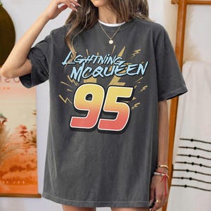 May include: A charcoal grey short-sleeve t-shirt featuring the text "LIGHTNING MCQUEEN" in light blue letters above a large, bold number "95" in a yellow-to-red gradient. The design includes yellow lightning bolt graphics on a dark background.