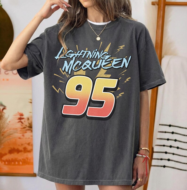 May include: A charcoal grey short-sleeve t-shirt featuring the text "LIGHTNING MCQUEEN" in light blue letters above a large, bold number "95" in a yellow-to-red gradient. The design includes yellow lightning bolt graphics on a dark background.