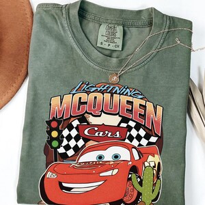 May include: An olive green crew neck t-shirt featuring a graphic print of the character Lightning McQueen from the movie Cars. The design includes a traffic light, checkered flags, and a cactus. The shirt has a Comfort Colors brand label.