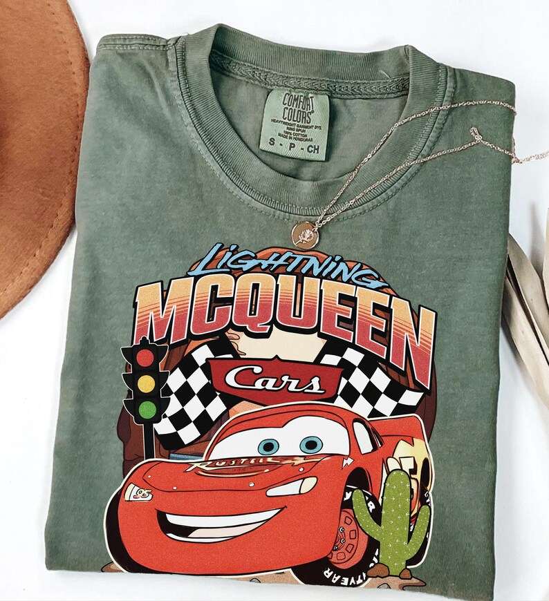 May include: An olive green crew neck t-shirt featuring a graphic print of the character Lightning McQueen from the movie Cars. The design includes a traffic light, checkered flags, and a cactus. The shirt has a Comfort Colors brand label.