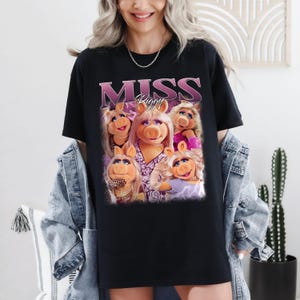 May include: Black t-shirt featuring a graphic of Miss Piggy and the text "MISS Piggy" in a pink and purple design. The graphic includes multiple images of Miss Piggy. The shirt is a casual style.