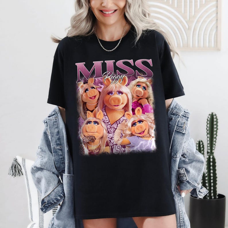 May include: Black t-shirt featuring a graphic of Miss Piggy and the text "MISS Piggy" in a pink and purple design. The graphic includes multiple images of Miss Piggy. The shirt is a casual style.