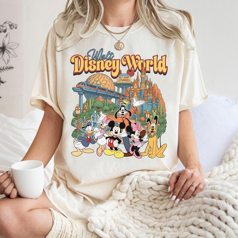 Pode incluir: Uma t-shirt de cor creme com uma impress&atilde;o gr&aacute;fica do Walt Disney World com o Mickey Mouse, a Minnie Mouse, o Pato Donald, a Margarida, o Pateta e o Pluto. O design inclui o castelo ic&oacute;nico, um monotrilho e o texto Walt Disney World.