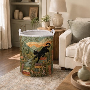 May include: A cylindrical fabric laundry hamper featuring a quilted texture and a printed design of a black cat climbing a stack of books in a forest setting with mushrooms, flowers, and a stream, finished with two white fabric handles.