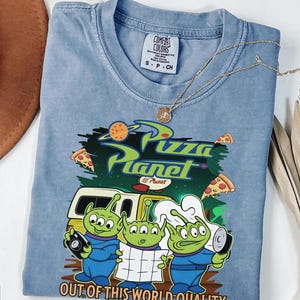May include: A blue short-sleeved t-shirt featuring a graphic of three green three-eyed aliens, a yellow vehicle, and pizza slices. The text reads Pizza Planet and Out of This World Quality. The shirt has a Comfort Colors label in the neck area.