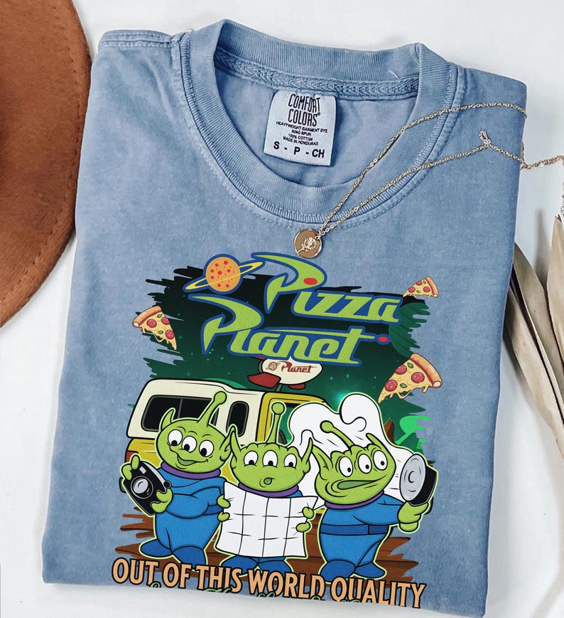 May include: A blue short-sleeved t-shirt featuring a graphic of three green three-eyed aliens, a yellow vehicle, and pizza slices. The text reads Pizza Planet and Out of This World Quality. The shirt has a Comfort Colors label in the neck area.