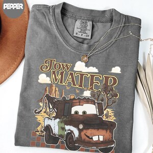 May include: A charcoal gray t-shirt featuring a graphic of the character Tow Mater from the movie Cars. The design includes desert scenery with cacti, rock formations, and the text Tow Mater in a retro font. The shirt has a Comfort Colors brand label.