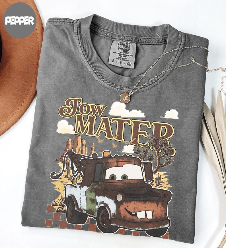 May include: A charcoal gray t-shirt featuring a graphic of the character Tow Mater from the movie Cars. The design includes desert scenery with cacti, rock formations, and the text Tow Mater in a retro font. The shirt has a Comfort Colors brand label.