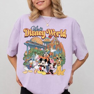 Pode incluir: Uma t-shirt roxo-claro com uma impress&atilde;o gr&aacute;fica do Walt Disney World e o texto Walt Disney World no topo. A ilustra&ccedil;&atilde;o inclui o Mickey Mouse, a Minnie Mouse, o Pato Donald, a Margarida, o Pateta e o Pluto em frente ao parque.