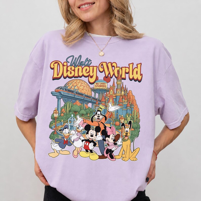 Pode incluir: Uma t-shirt roxo-claro com uma impress&atilde;o gr&aacute;fica do Walt Disney World e o texto Walt Disney World no topo. A ilustra&ccedil;&atilde;o inclui o Mickey Mouse, a Minnie Mouse, o Pato Donald, a Margarida, o Pateta e o Pluto em frente ao parque.