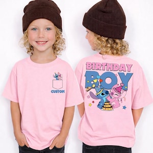 May include: Pink short-sleeved t-shirt with a pocket design featuring cartoon characters. The back of the shirt says "BIRTHDAY BOY" with cartoon characters and a cake. The word "CUSTOM" is printed on the pocket.